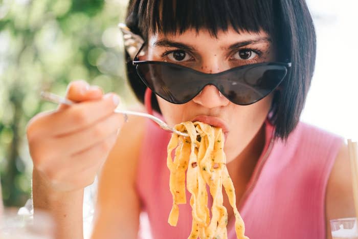 A woman with short dark hair, wearing sunglasses and a sleeveless top, eats pasta while looking at the camera