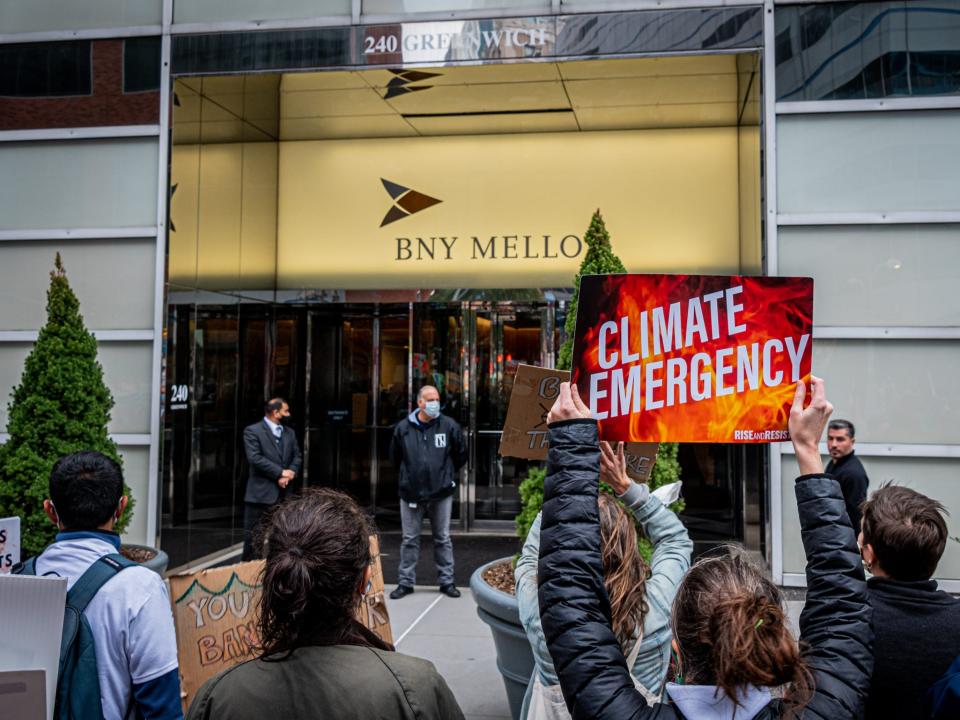 A bright red and orange poster that says "climate emergency" in front of a golden BNY Mellon sign on a building.