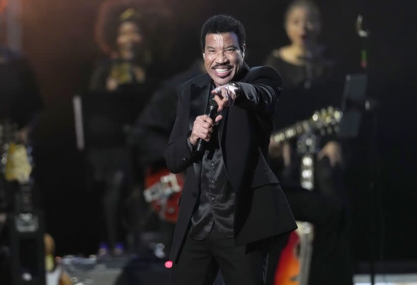 Lionel Richie is wearing a black suit and is pointing while performing on stage with a microphone held to his face