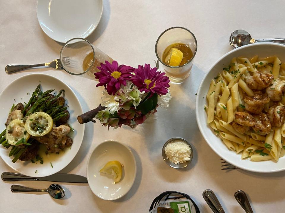 A flat lay angle of the table with a chicken, lemon, and asparagus plate and a shrimp and pasta bowl surrounded by utensils, iced tea glasses, and a small bouquet of flowers.