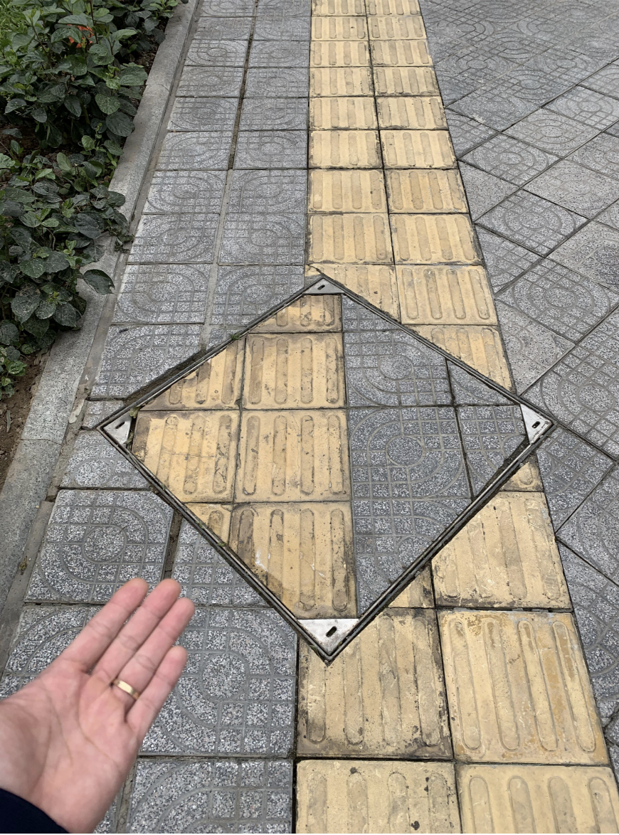 A person's hand pointing to a path with the sewer cap that is lined up incorrectly with the sidewalk