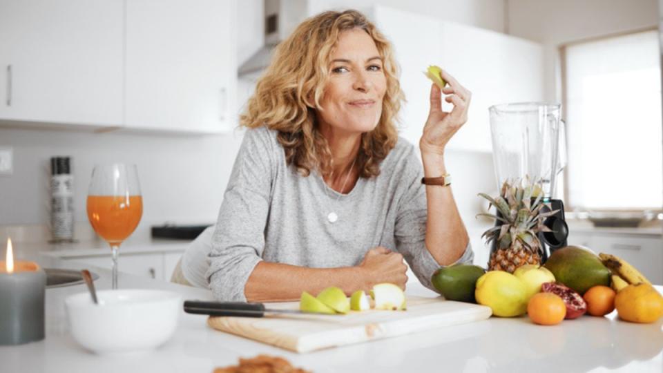 Mature woman at her kitchen counter eating an apple, which is one of the best healthy eating hacks, beside other fresh fruit