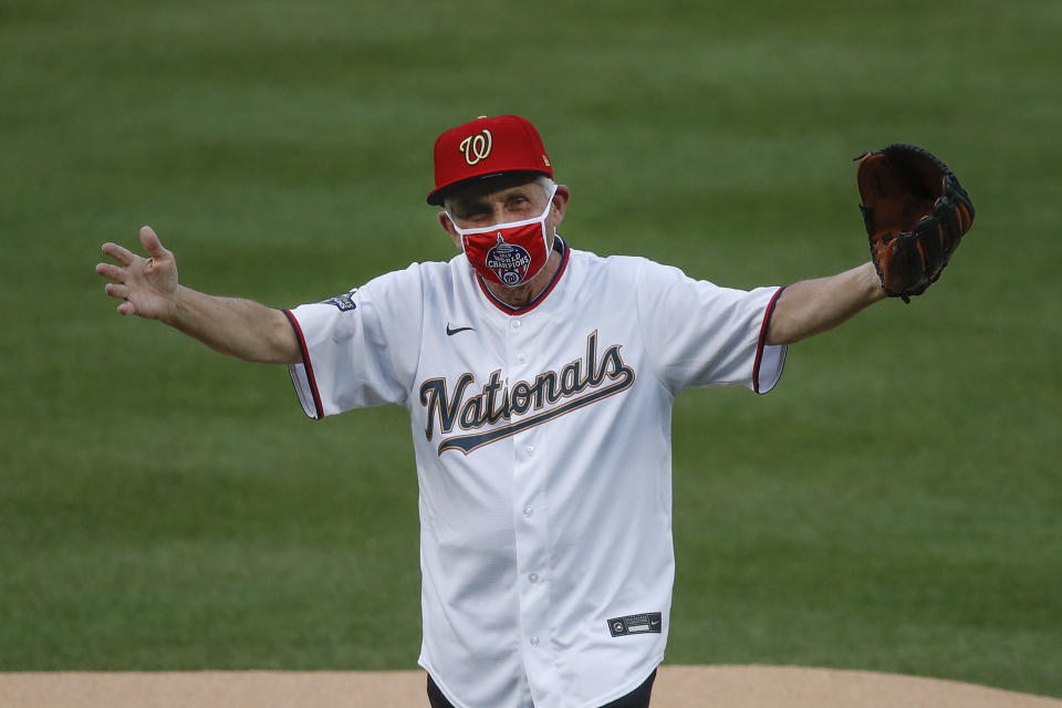 Dr. Anthony Fauci in a Washington Nationals jersey, mask and hat with a baseball glove raising his hands.