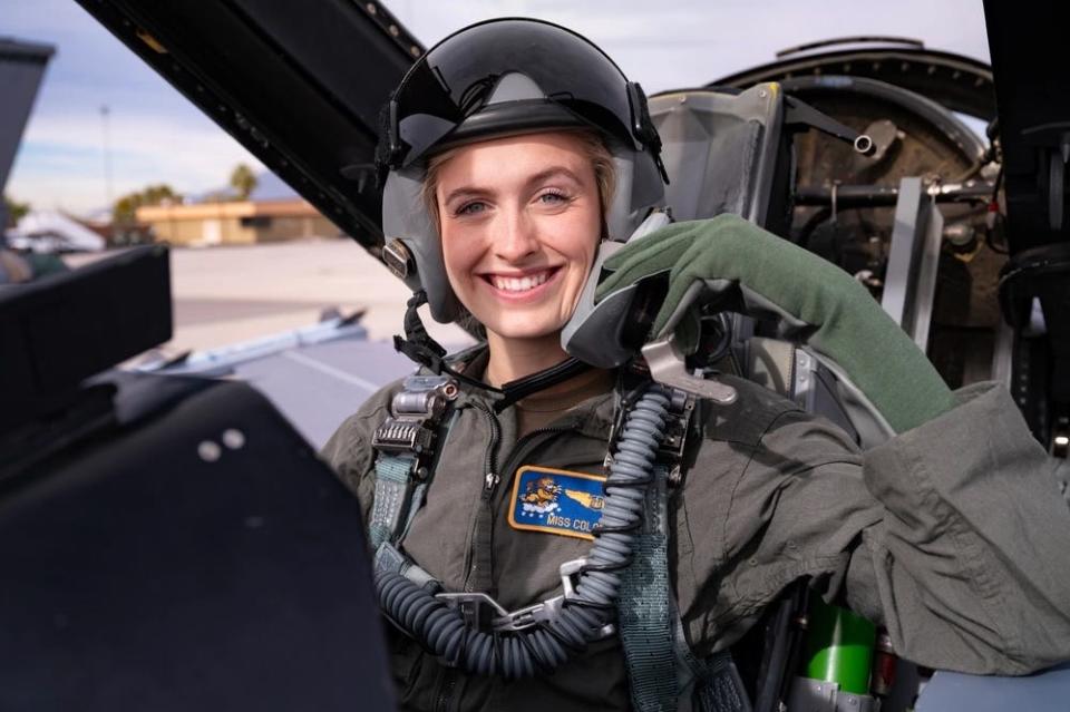 A woman in a flight suit and a helmet smiles for a photo from the cockpit of an aircraft