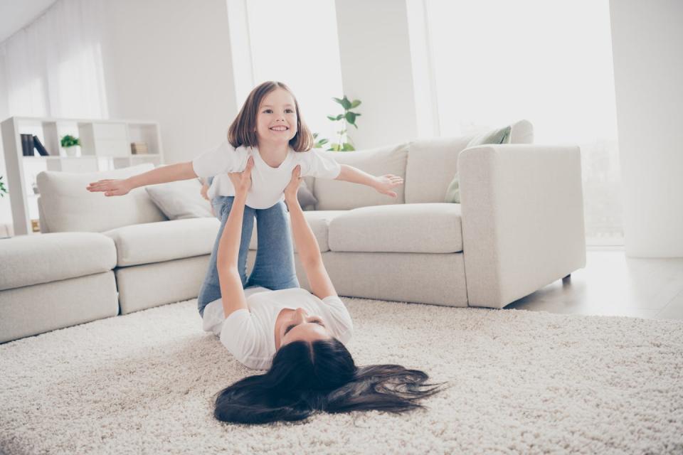 A woman lying on her back on a white carpet is holding a little girl who is pretending to fly.  There is a white sofa behind them.