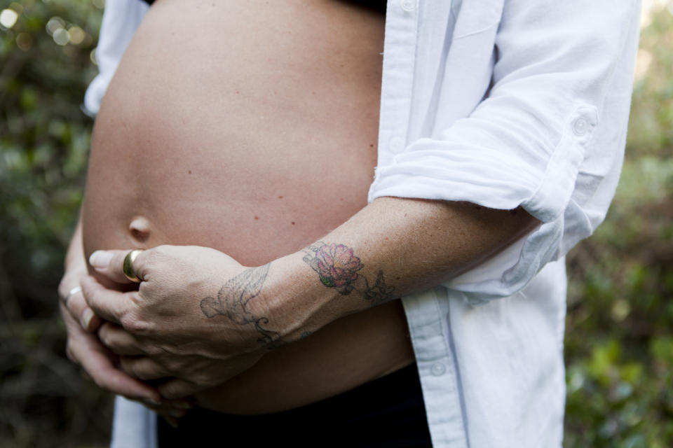 Close up shot of a woman's pregnant belly. Her blue shirt is unbuttoned and her hands are cupping her belly. She has coloured floral tattoos on her hand.