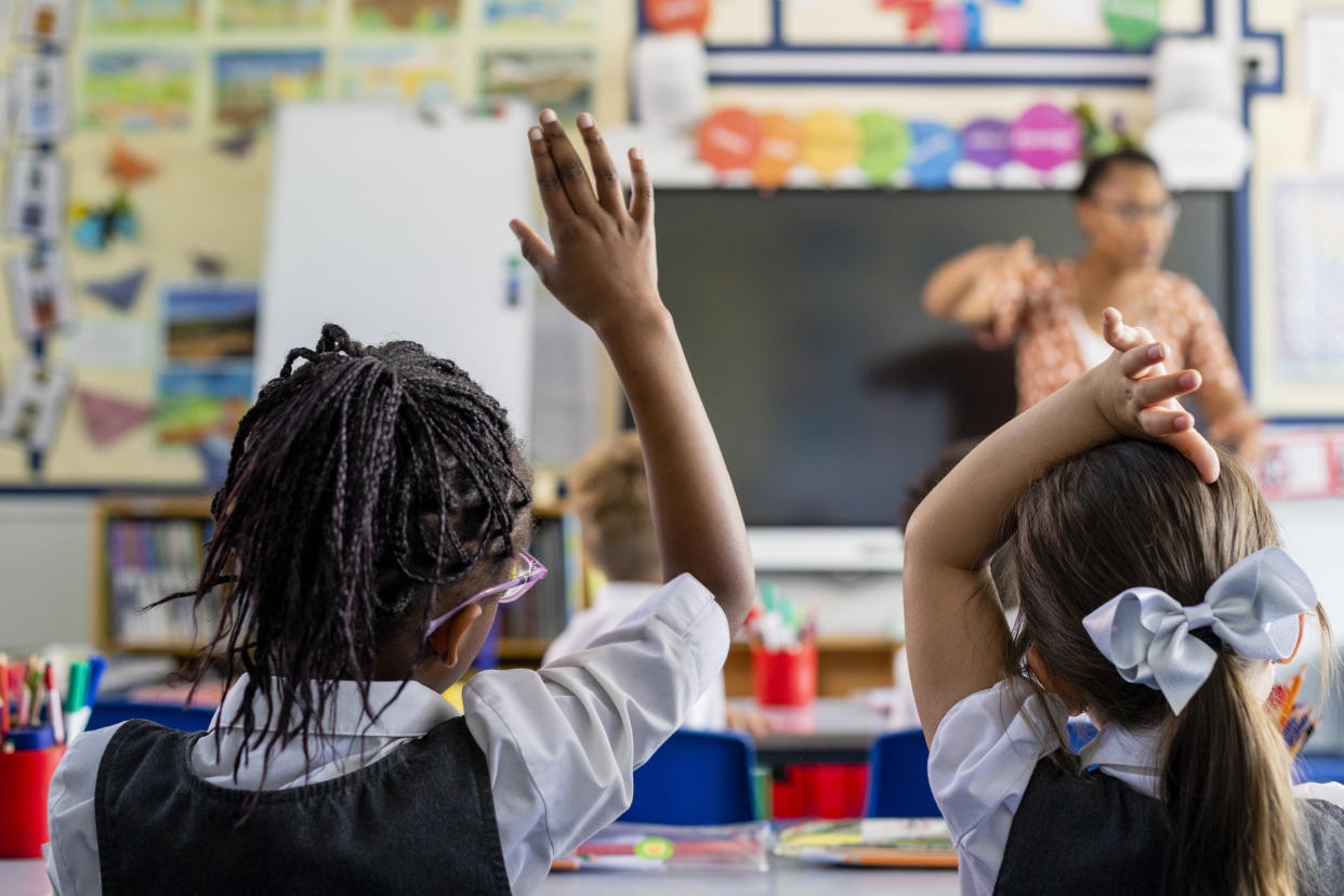 Primary school students sitting in a classroom being taught by a teacher in the North East of England. The students are answering questions with their arms in the air.