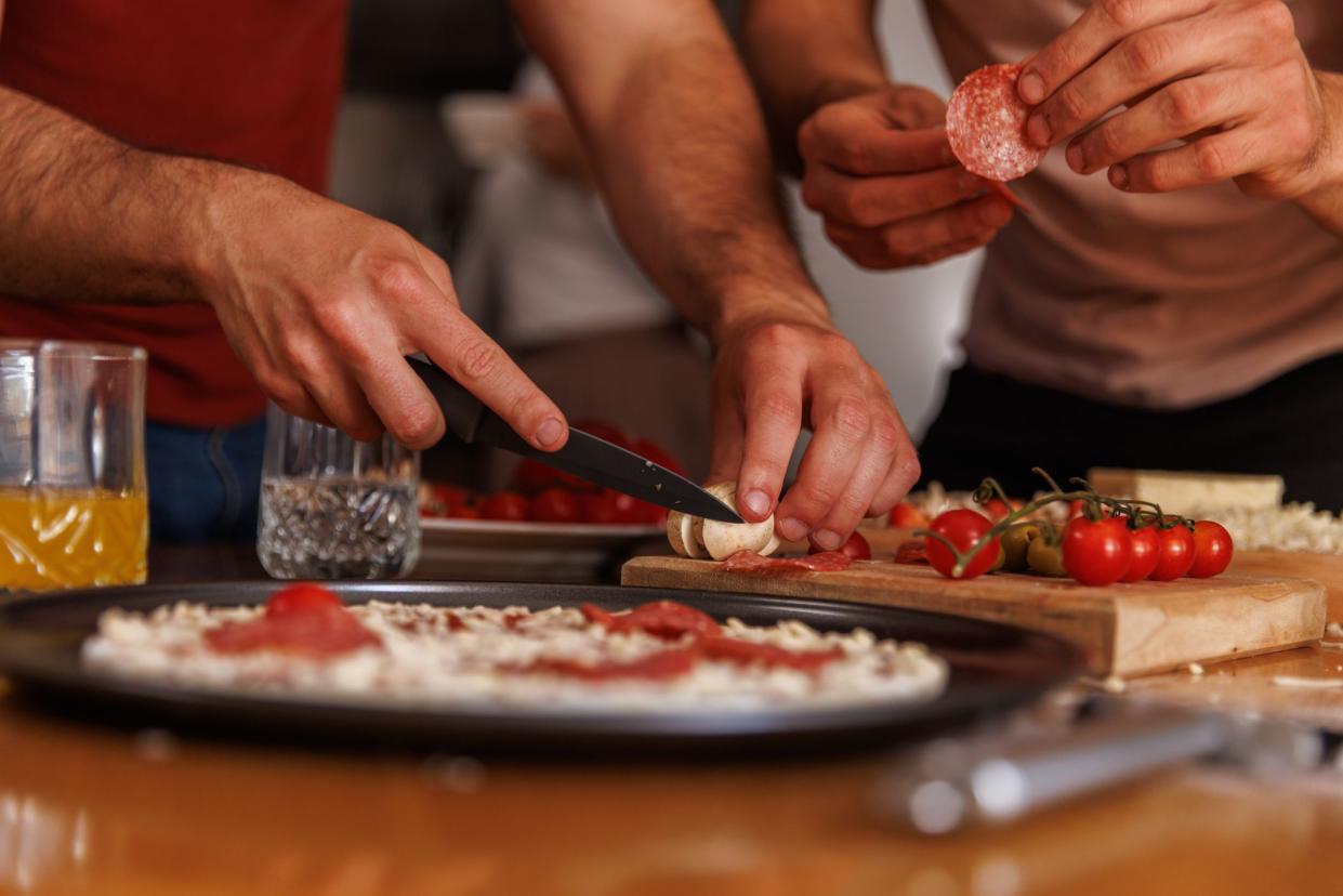 Candid shot of small group of male friends putting toppings on a homemade pizza that they are making.