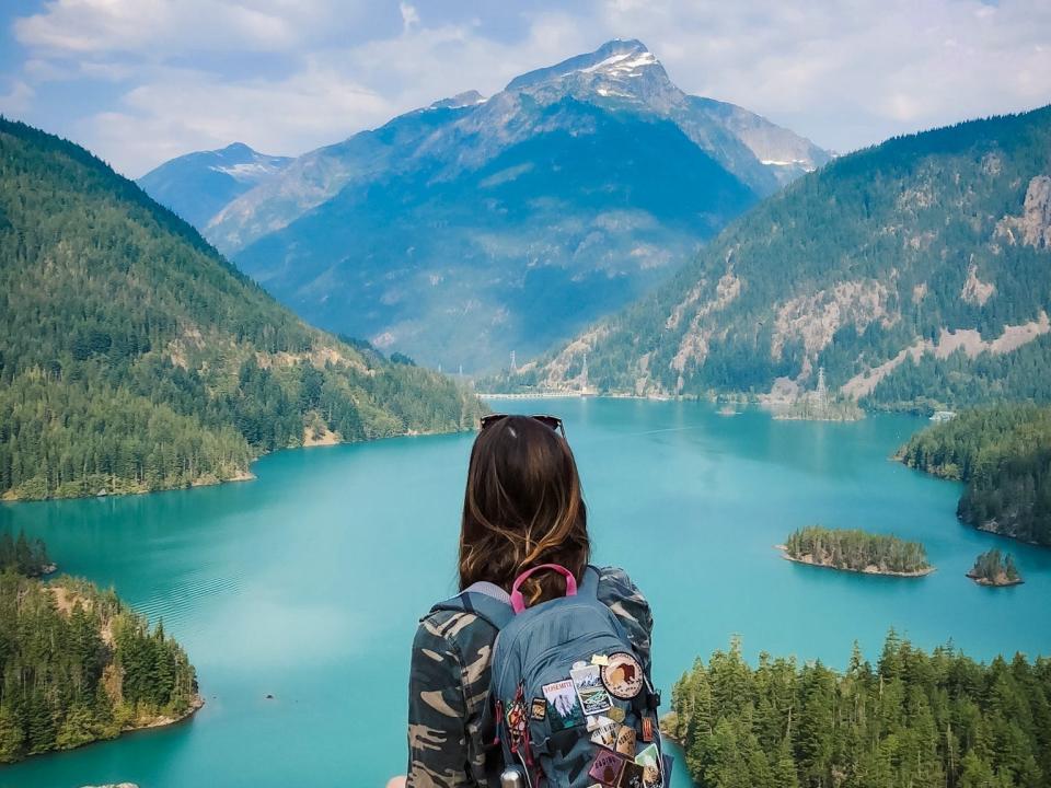 Emily sits on a rock wearing a backpack covered in patches. She looks out at stunning blue water, mountains, and trees. 