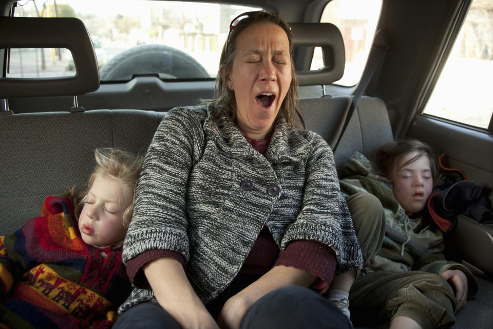 A woman yawns while sitting in a car's backseat with two sleeping children beside her. The children are dressed in colorful and cozy clothing