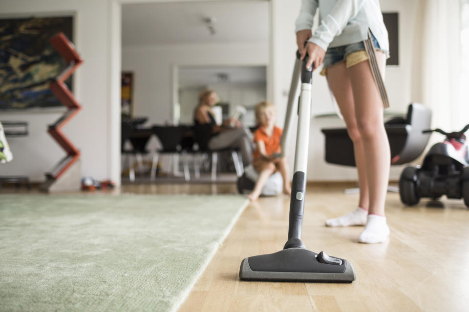 A kid vacuums in their family's house while their sibling and mom sit at a distance form them