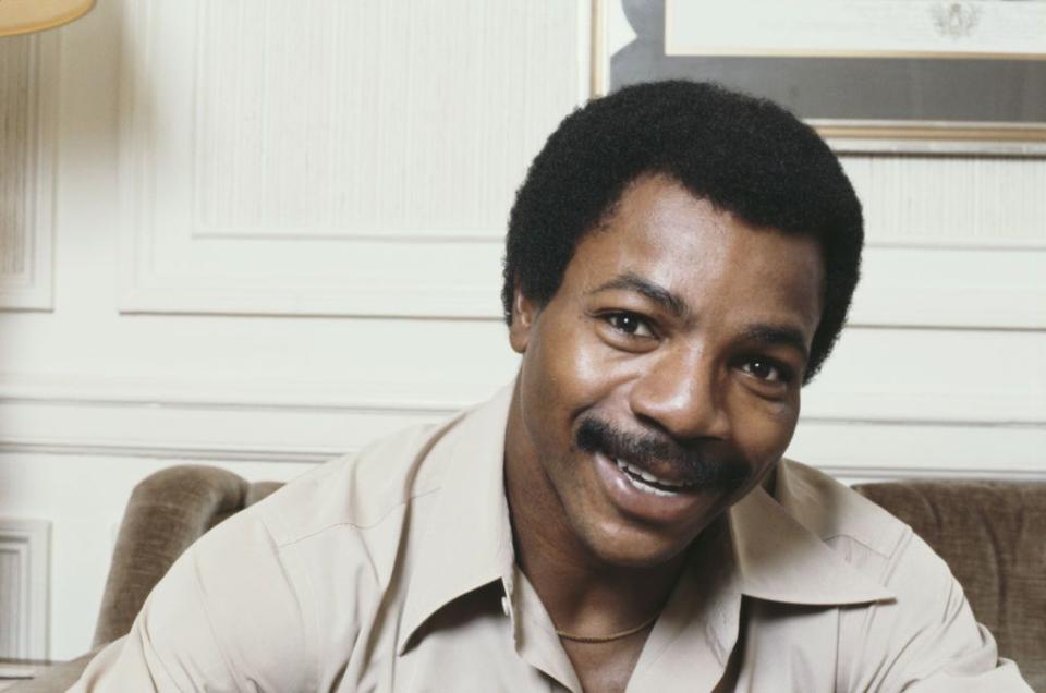 carl weathers smiles at the camera, he wears a brown collared shirt and sits on a couch, behind him a painting hangs on the wall