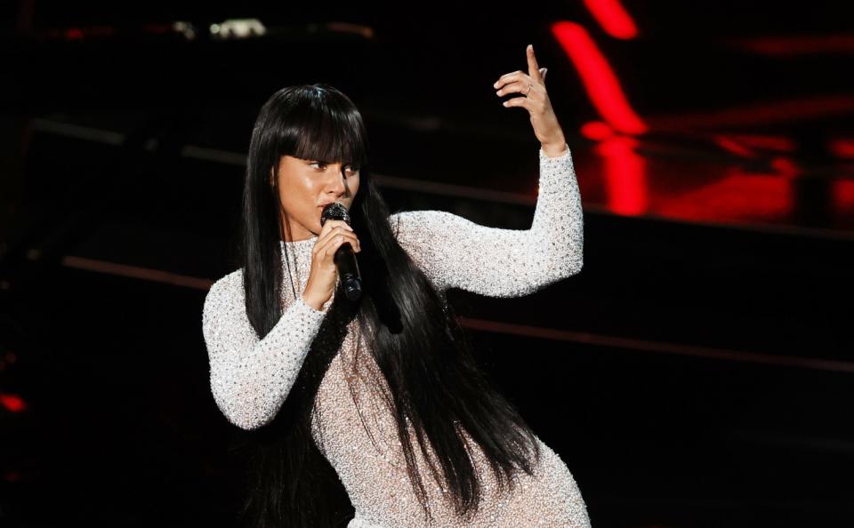 Alicia Keys, in a form-fitting white sequined gown, sings into the microphone.