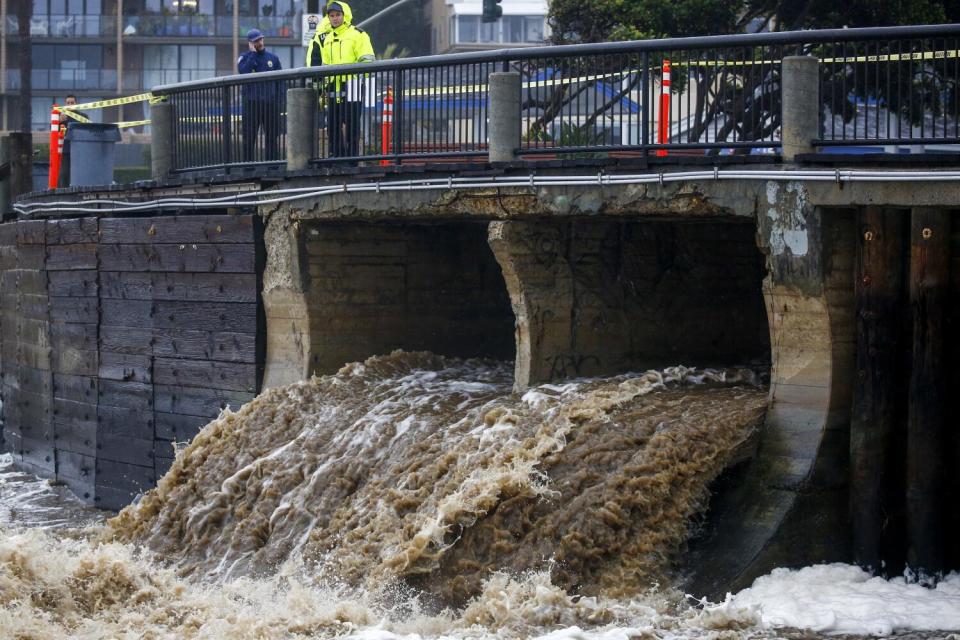 Stormwater pours into the ocean from an outfall channel as bystanders look on.