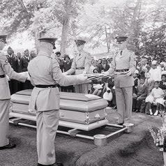 A military honor guard folds an American flag over the casket of Medgar Evers, the slain civil rights leader. Widow Myrlie B. Evers, and two of her three children, Darrell Kenyetta, 9, and Reena Denise, 7, are shown seated at extreme right