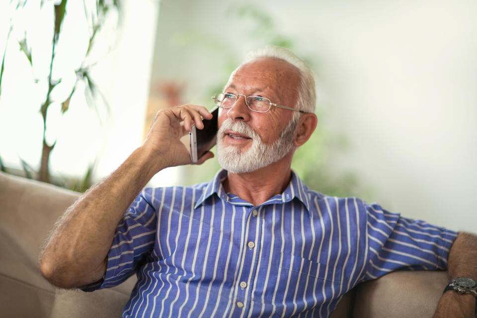 Seated senior man with beard in a blue shirt with white stripes, talking on mobile phone.