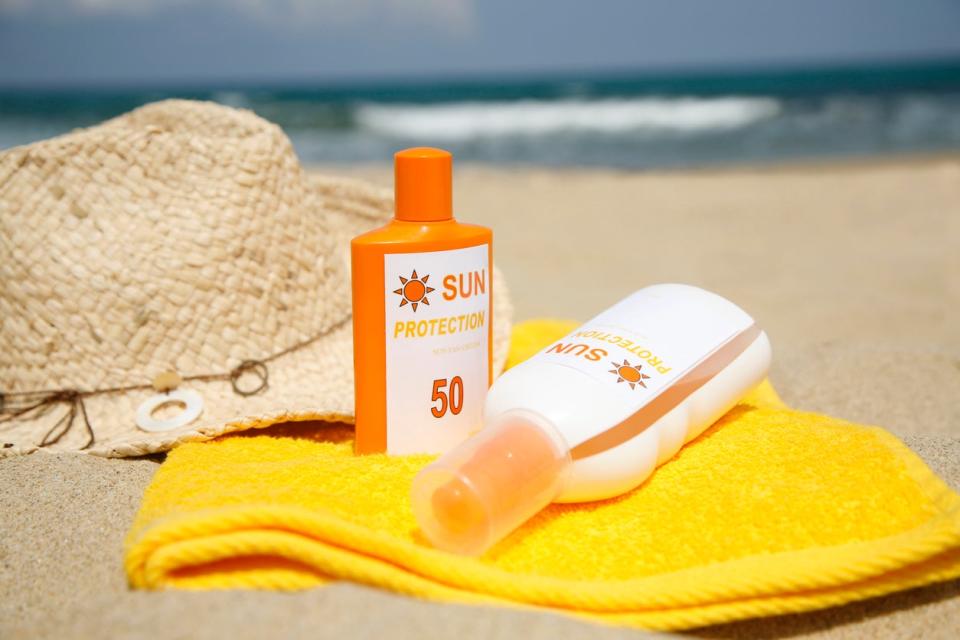 Two orange and white bottles of sunscreen on top of yellow beach towel in front of straw sun hat on sand