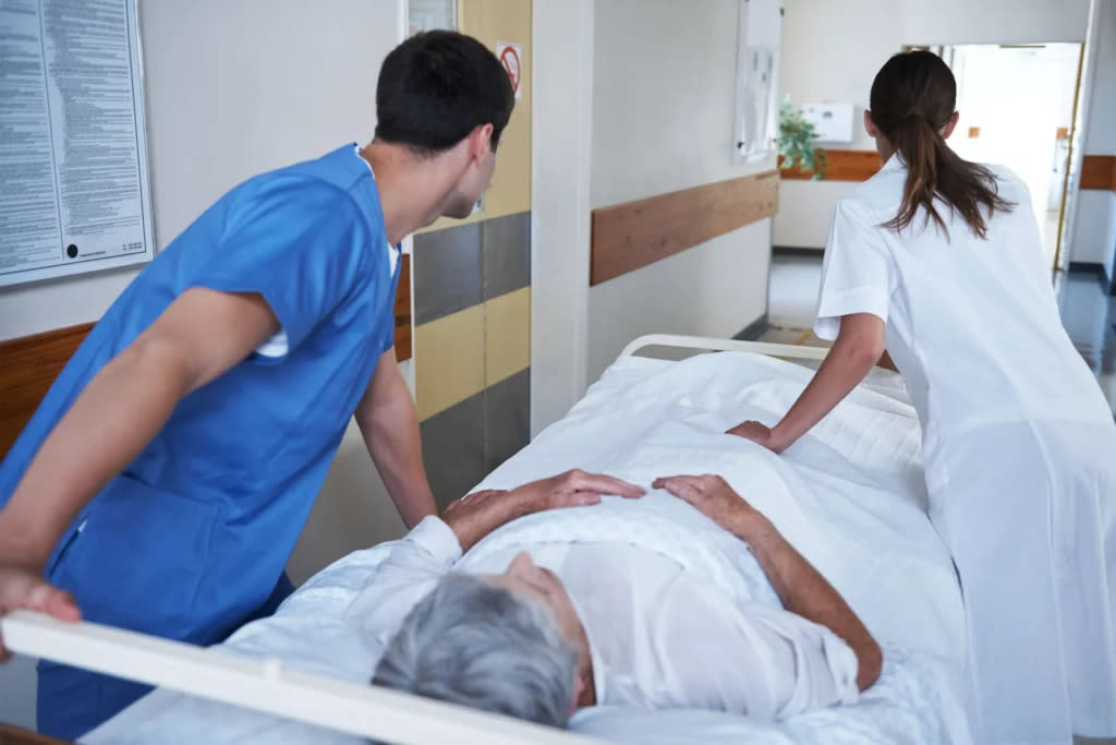 A male orderly in medical scrubs and a female nurse wheel an elderly patient on a gurney through a hospital corridor.