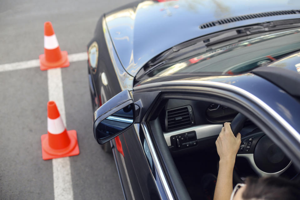 Person in a car at a driving test with instructor holding door and traffic cone visible