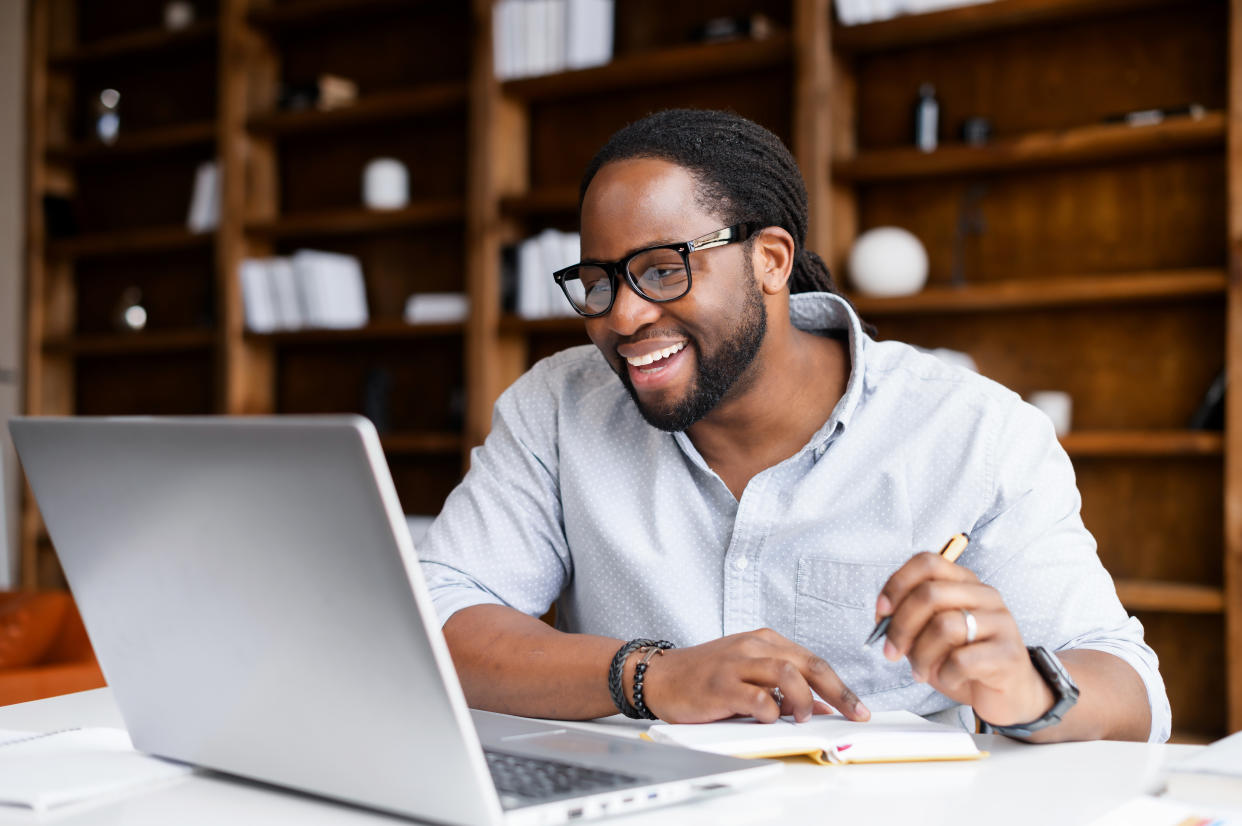 Smiling African-American guy takes notes watching webinars on the laptop,researching business tasks, a male student is studying online, listening video lectures, writes down, e-learning concept