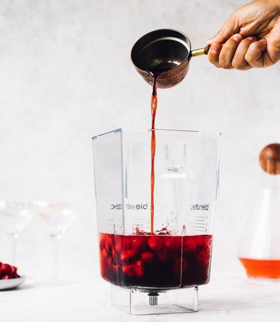 A hand is pouring cherry juice from a measuring cup into a blender filled with cherries. Two cocktails and a decanter are in the background