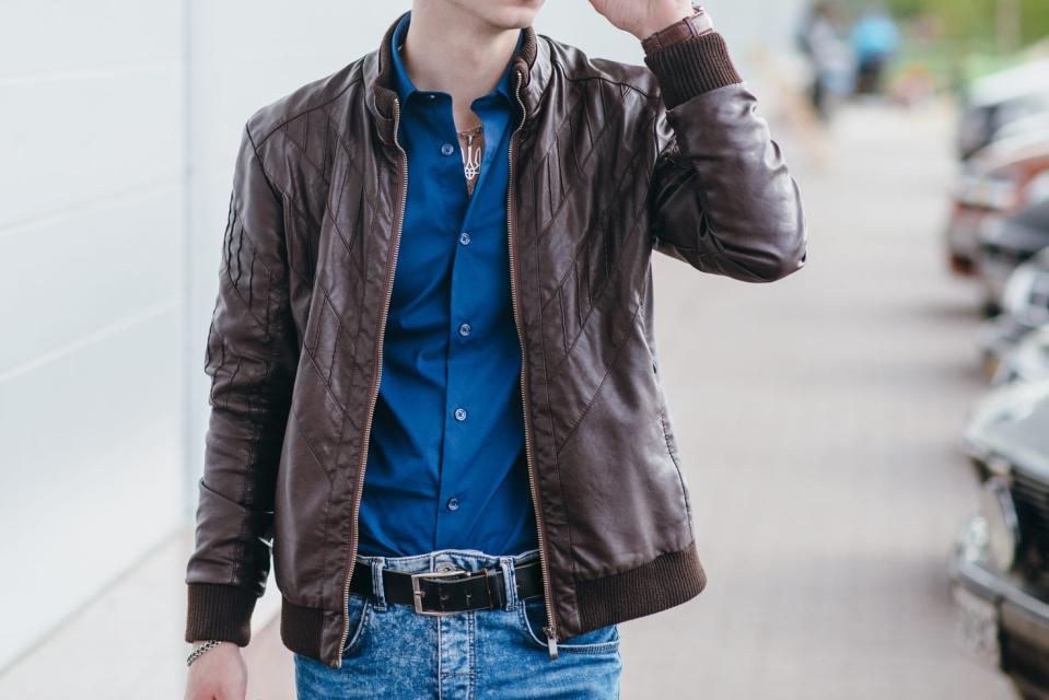 A man wears a brown leather jacket over a blue button-down shirt with jeans while walking down a city street, Parked cars line the sidewalk.