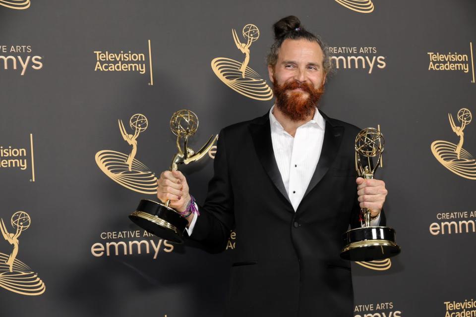 white lotus composer cristobal tapia de veer holding up two emmy awards, wearing a black suit with a medium beard and a man bun