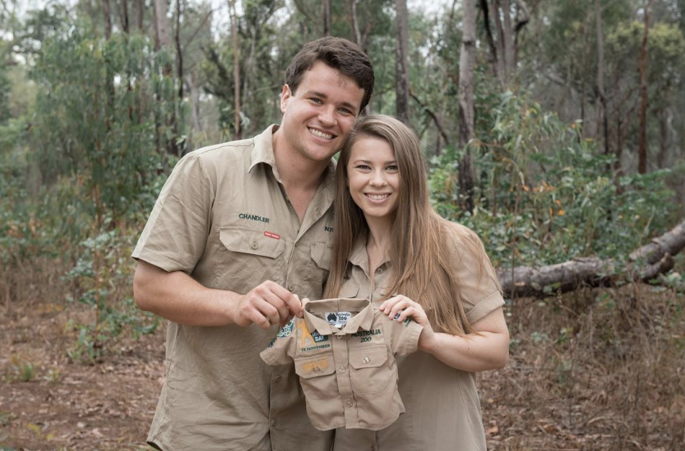 Bindi Irwin and Chandler Powell holding up a baby zookeeper outfit