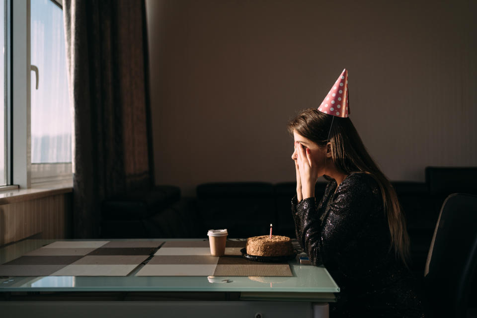 A woman in a party hat sits alone at a table with a small cake and a lit candle in front of her, covering her face in her hands