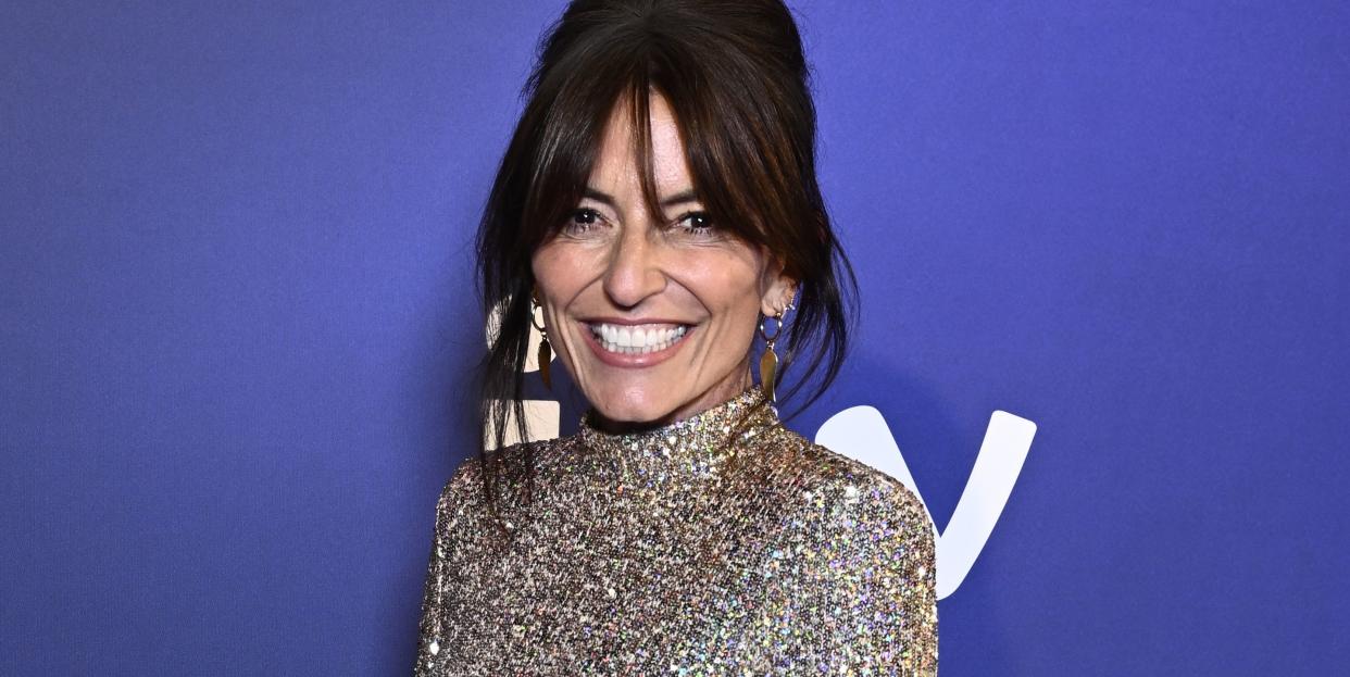 davina mccall, a woman stands smiling at the camera, she has dark hair worn up with a fringe, she wears a short silver sequin dress with long sleeves