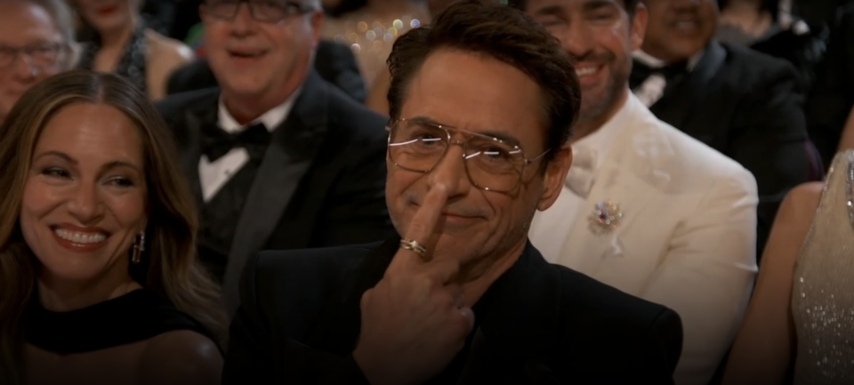 Man in glasses giving a peace sign, seated at an event with attendees in formal wear