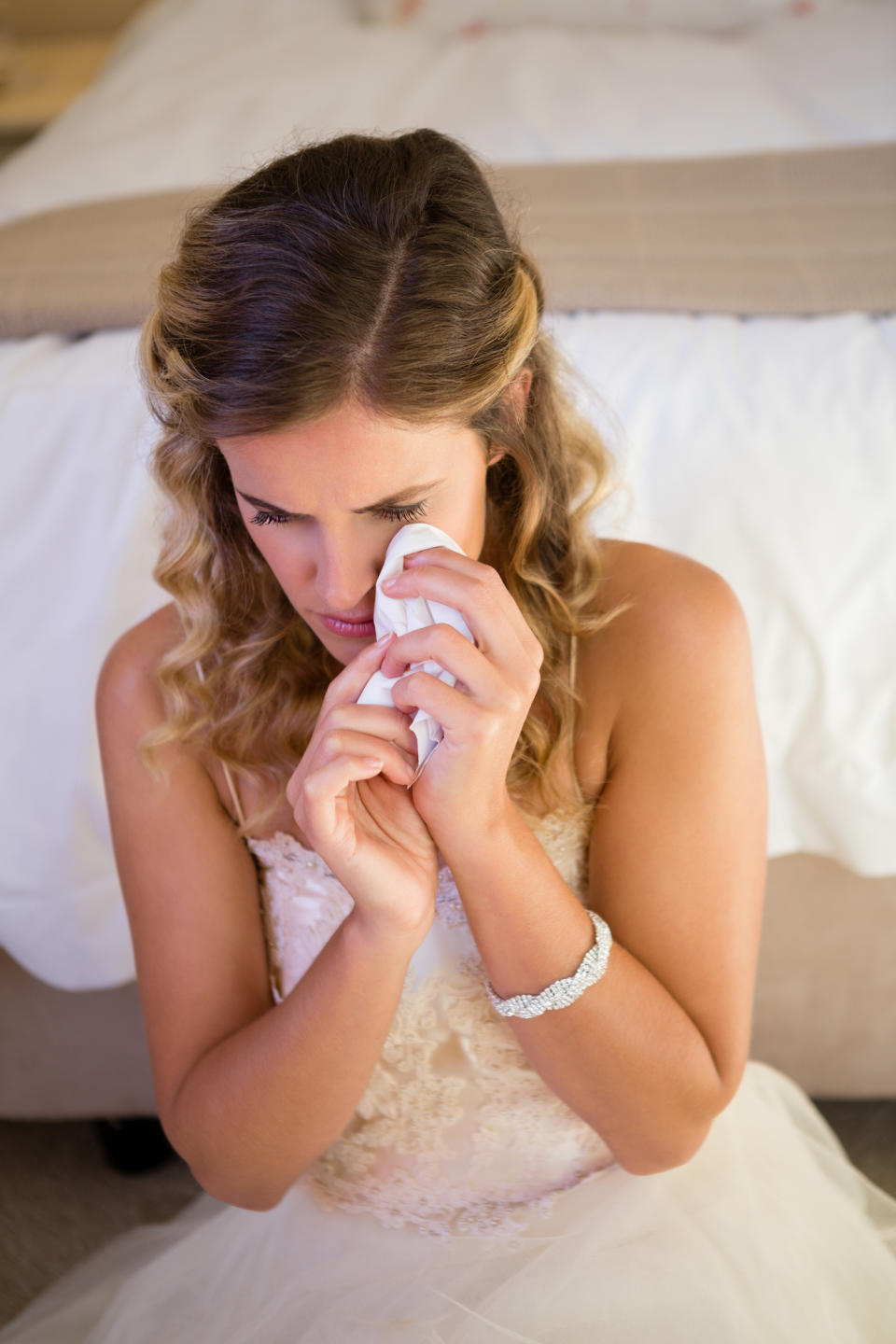 A bride in a wedding dress sits on a bed while wiping away her tears with a tissue, appearing emotional