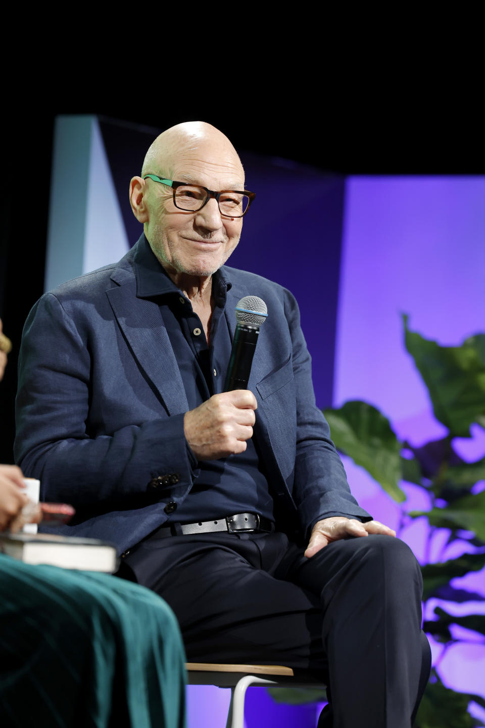Patrick Stewart in a blazer with a microphone seated on stage, smiling