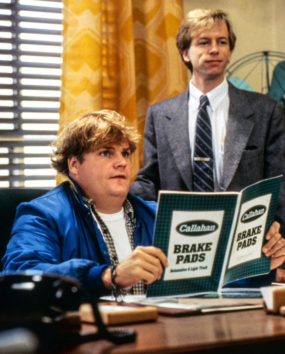 Chris Farley in Tommy Boy sitting at a desk holding a brochure about brake pads