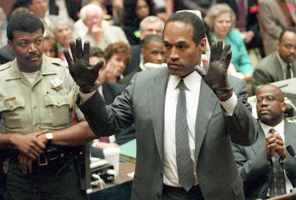 oj simpson raises both of his hands in the air in front of him and looks left, he wears black gloves and a gray suit and tie with a collared white shirt, many people look on from the background