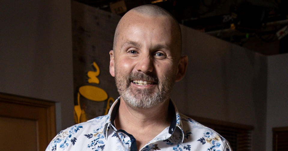 Neighbours star Ryan Moloney smiles at the camera wearing a white shirt with a blue floral pattern on it