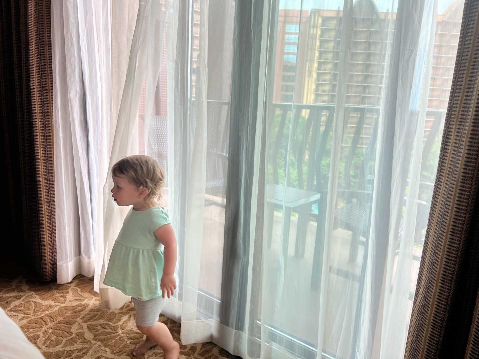 A young child standing in the curtains of large windows and door leading to a balcony.