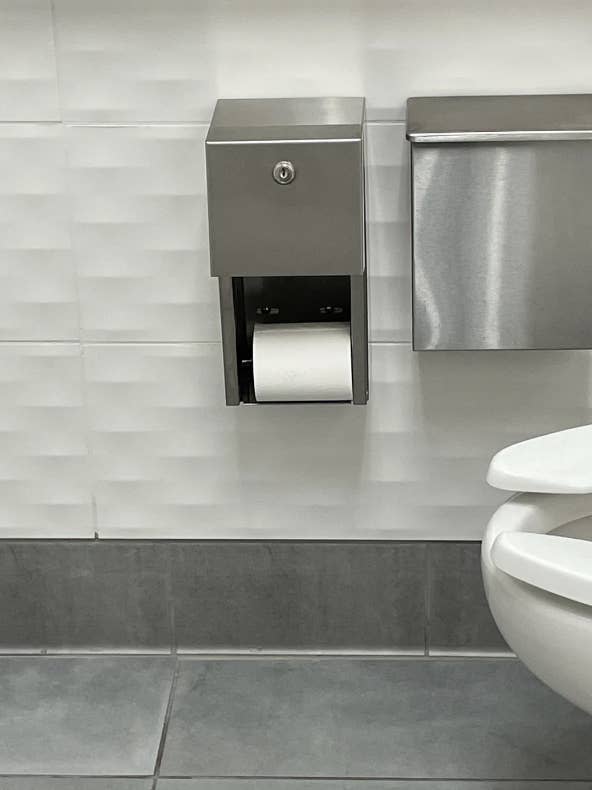 A modern bathroom with a stainless steel toilet paper dispenser, white textured tiles, and a partial view of a stainless steel toilet