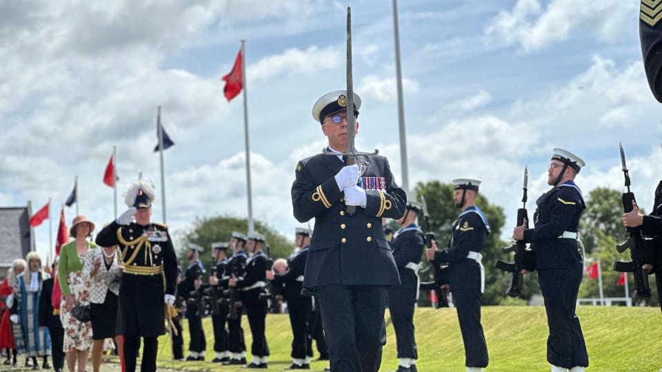 A man in a blue military uniform with colourful medals on his breast and a white cap and gloves carries the Sword of State along the ceremonial walkway. He is followed by the lieutenant governor, who is in full military uniform including a hat with white plumage and is saluting the Naval Guard of Honour which is lining the path.