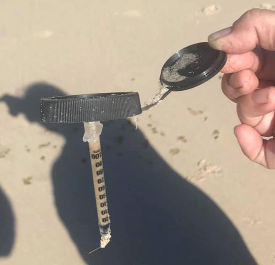 A needle is held by a woman with the plastic lid covered in sand at a beach.