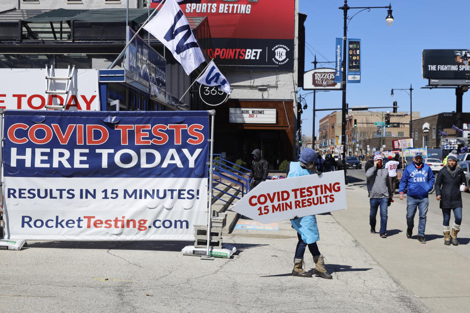 A person stands with a sign related to COVID-19 testing as Chicago Cubs fans pass by outside of Wrigley Field on the opening day baseball game between the Chicago Cubs and the Pittsburgh Pirates, Thursday, April 1, 2021 at Chicago. Fans are back at the ballpark after they were shut out during the regular season last year because of the coronavirus pandemic. (AP Photo/Shafkat Anowar)