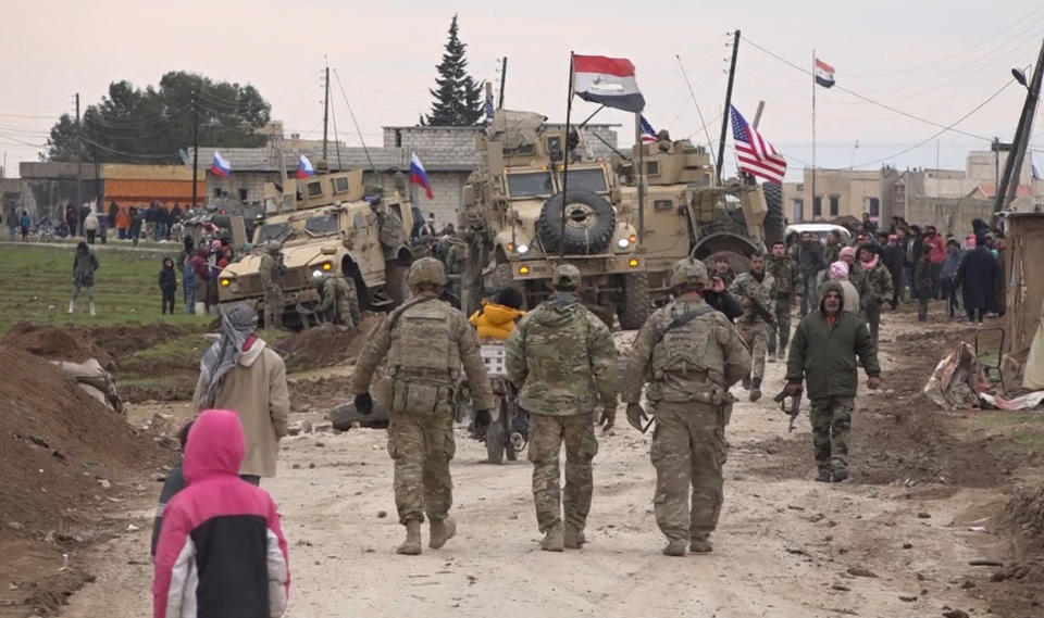 In this frame grab from video, Russian, Syrian and others gather next to an American military convoy stuck in the village of Khirbet Ammu, east of Qamishli city, Syria, Wednesday, Feb. 12, 2020. The Syrian official news agency SANA, said Wednesday, that locals had gathered at an army checkpoint, pelting the U.S. convoy with stones and taking down a U.S. flag flying on a vehicle when troops fired with live ammunition and smoke bombs. (AP Photo)