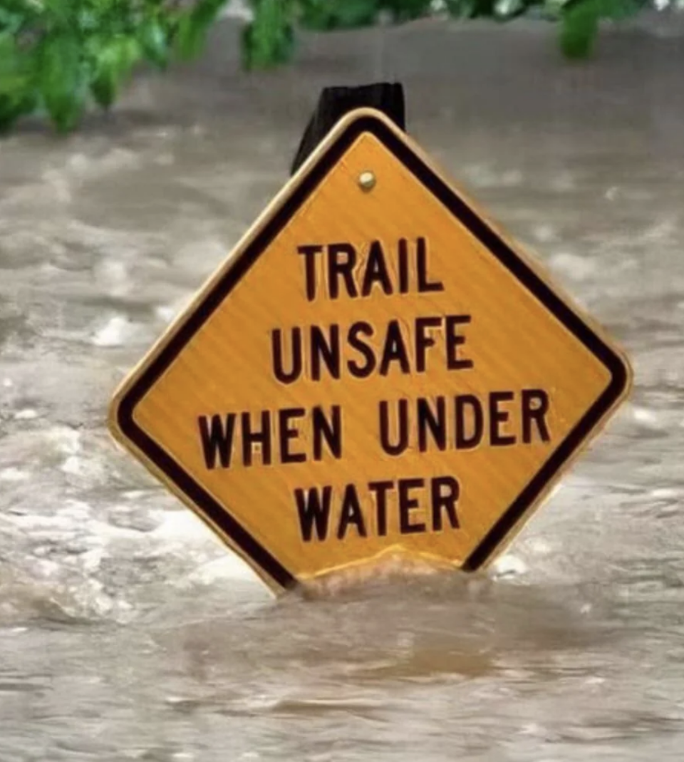 A sign in floodwater reads "Trail unsafe when under water," indicating danger on a submerged trail