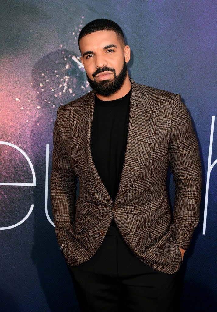 Drake stands posing in a plaid blazer, hands in pockets, at an event