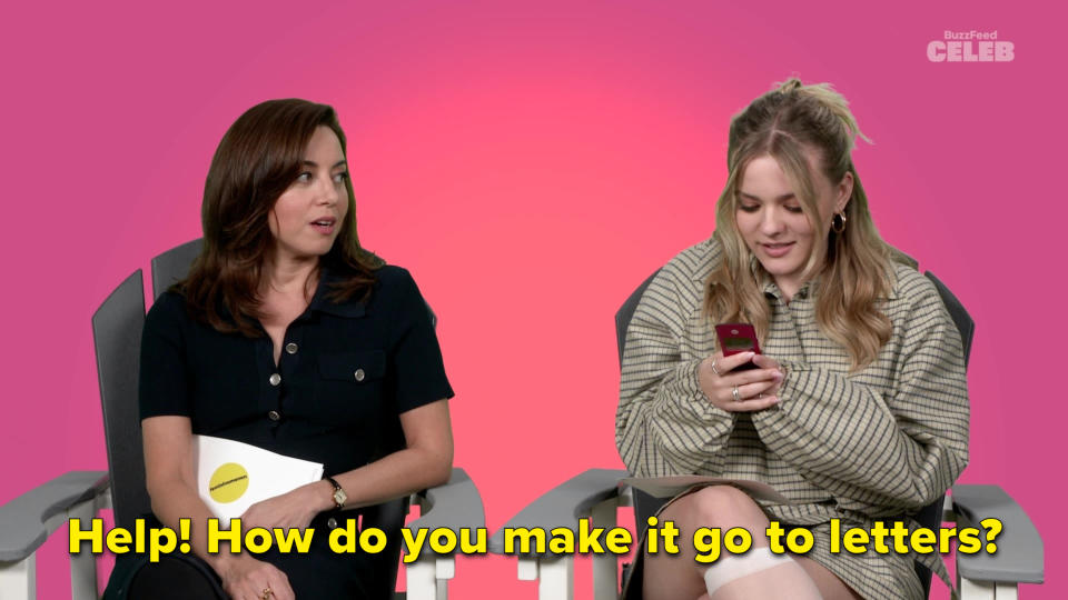 Aubrey Plaza, in a dark dress, and Maisy Stella, in a loose outfit, seated side by side. Aubrey holds a white notepad, Maisy looks at her phone. Background: BuzzFeed Celeb