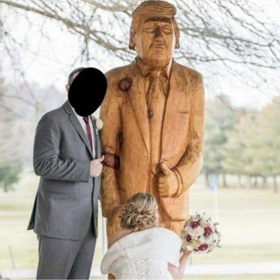 A bride kneeling in front of a wooden statue of Donald Trump while the groom looks