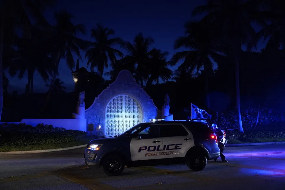 Police crouch behind a vehicle with Police Palm Beach painted on the side by a gated entrance flanked by a Trump flag beneath palm trees faintly illuminated by the night sky.