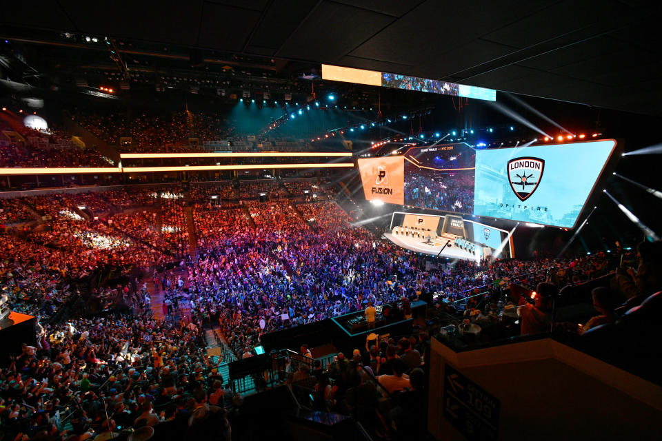 A sold out crowd in a large arena with a brightly lit stage with large screens on the floor of the arena during an esports event.