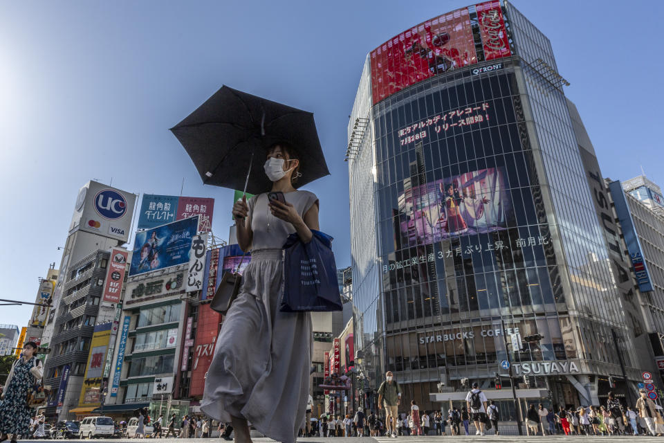 A woman wearing a mask and carrying an umbrella crosses a street with buildings covered with electronic advertisements and with signs saying, for example, Starbucks Coffee and Tsutaya.