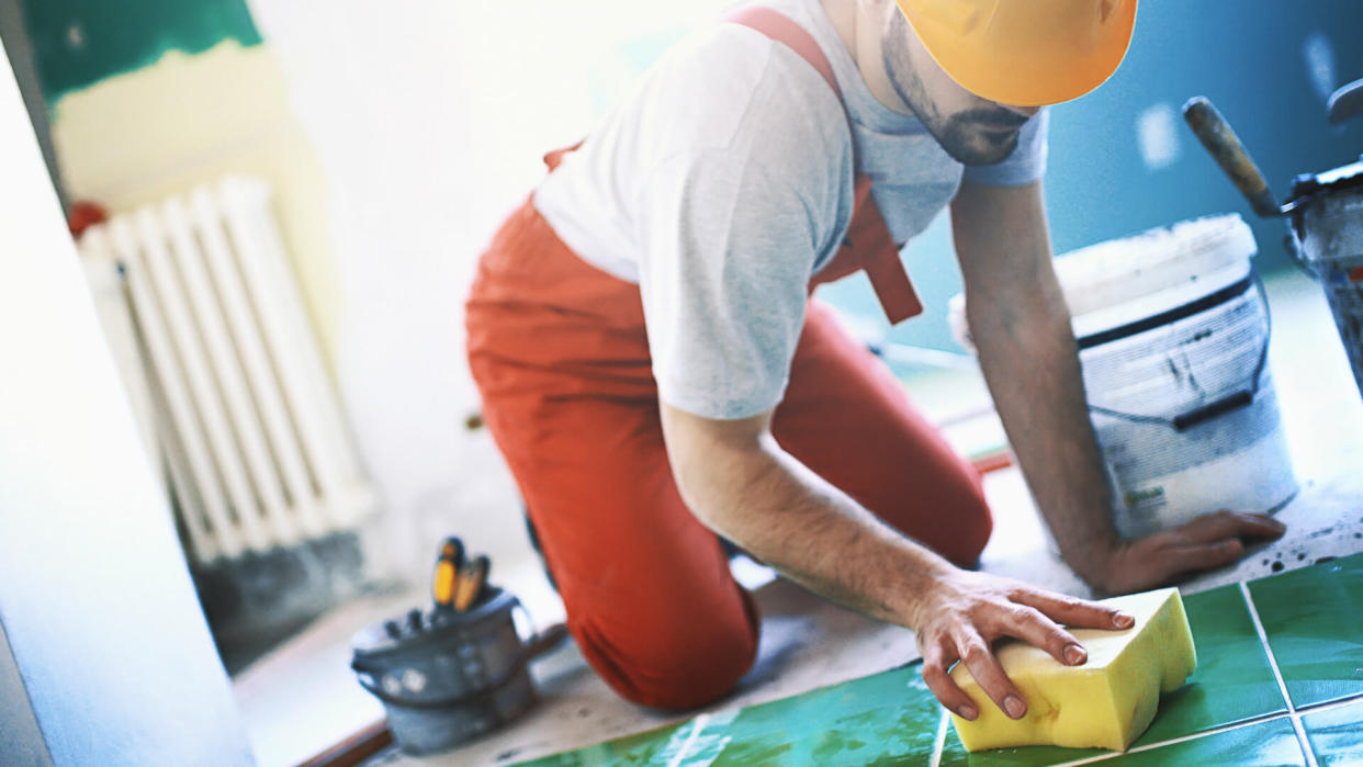 Closeup side view of a handyman installing green ceramic tiles over apartment floor.